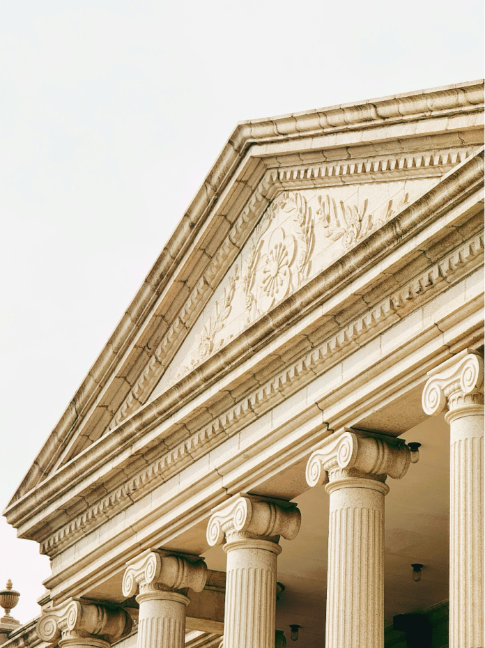 An ornate stone building facade with fluted columns, carved floral reliefs, and a neoclassical pediment rises against a bright sky.