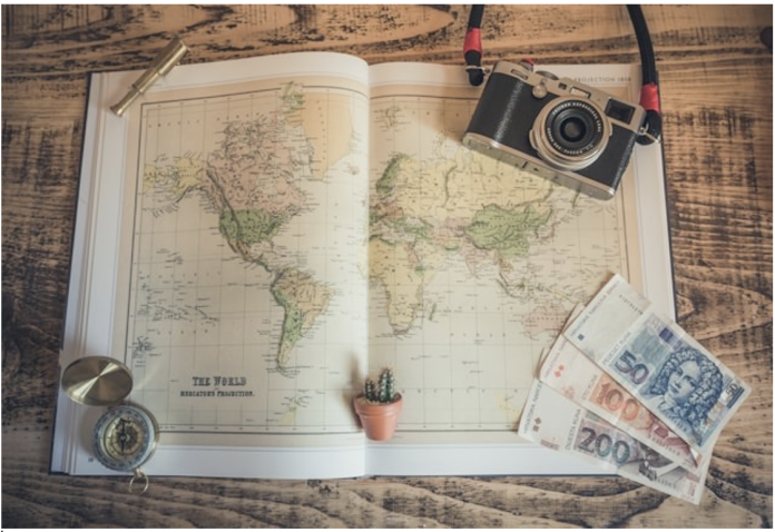 An open world atlas lies on a wooden table with a vintage camera, compass, euro banknotes, and a small cactus arranged for travel planning.