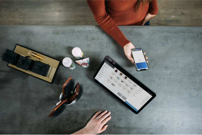 A rust-sweatered person holds a smartphone over a counter with a tablet, grooming items, and gift boxes.