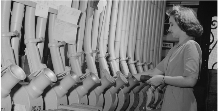 A woman in a dress operates a row of industrial control pipes and levers in a black and white archival setting.